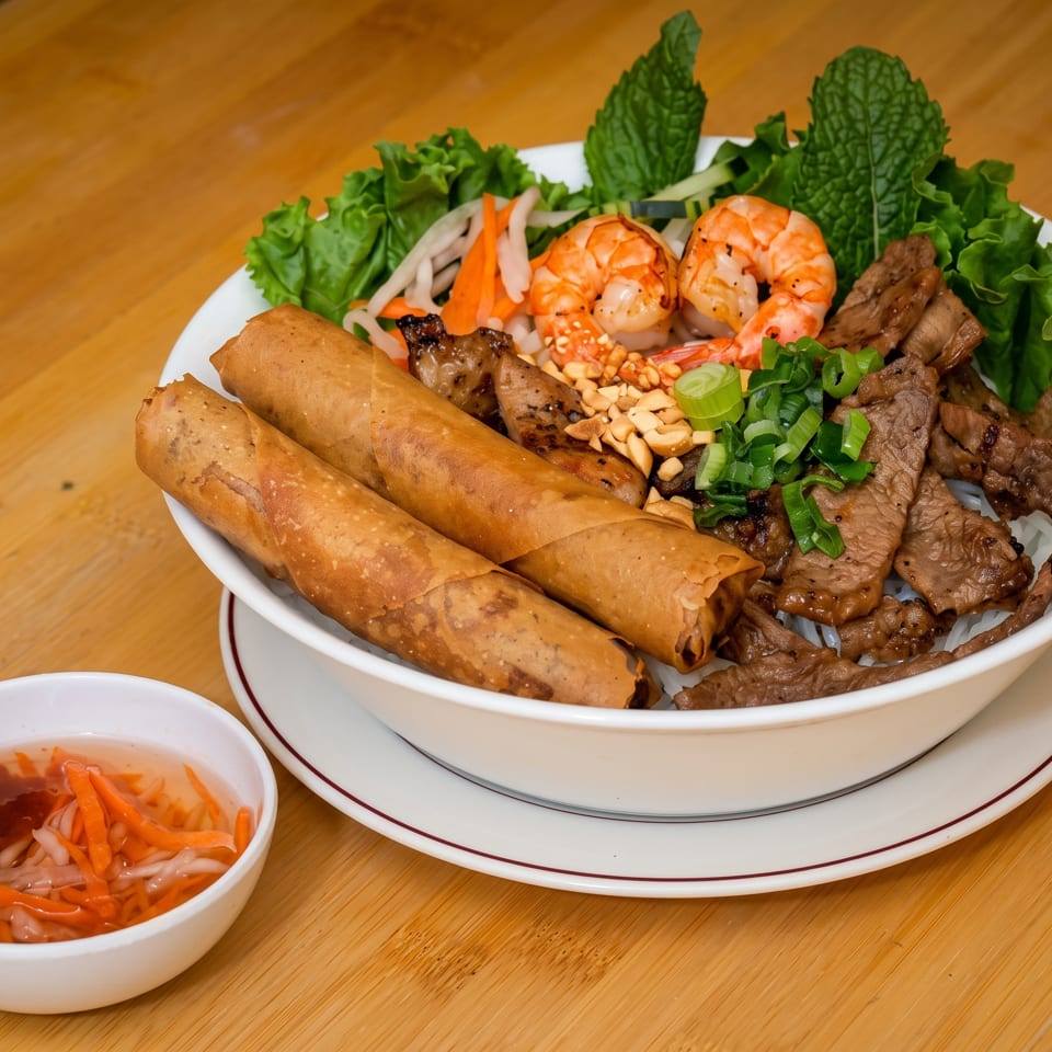A Vietnamese vermicelli bowl with grilled beef, shrimp, egg rolls, and a side of dipping sauce.