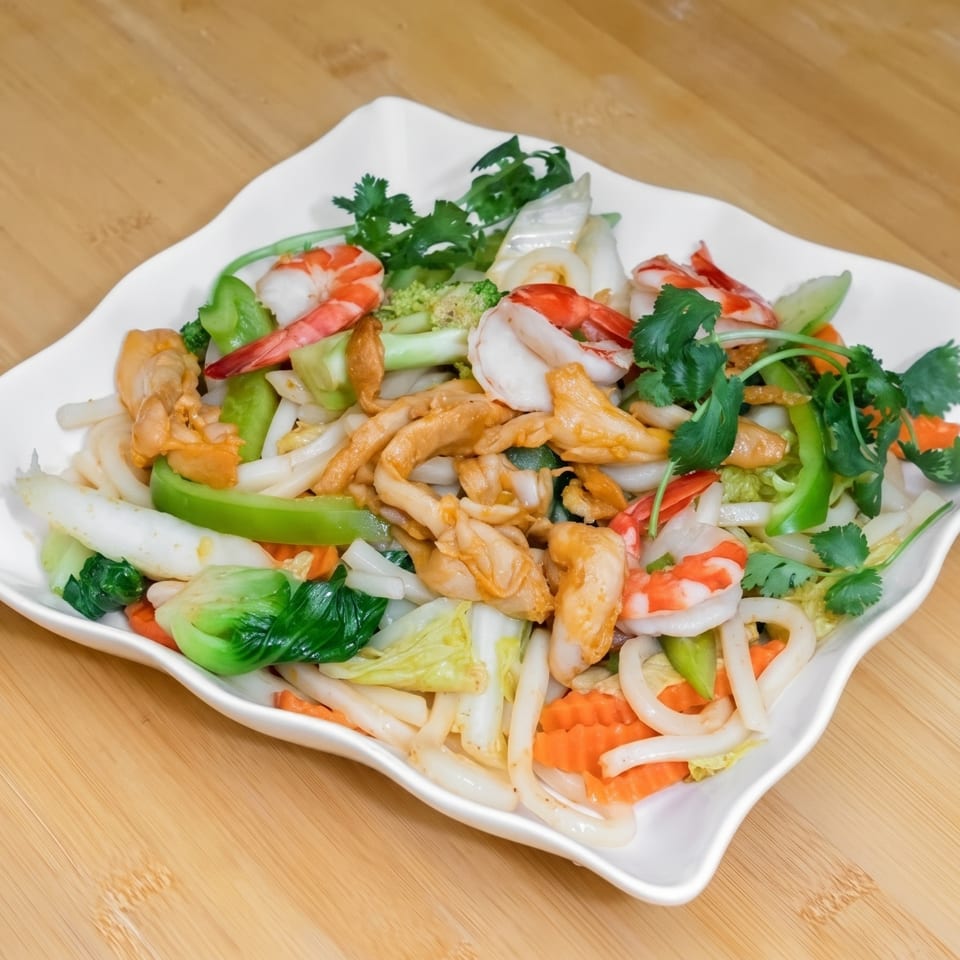 A white platter of stir-fried noodles with shrimp, chicken, and colorful vegetables on a wooden table.